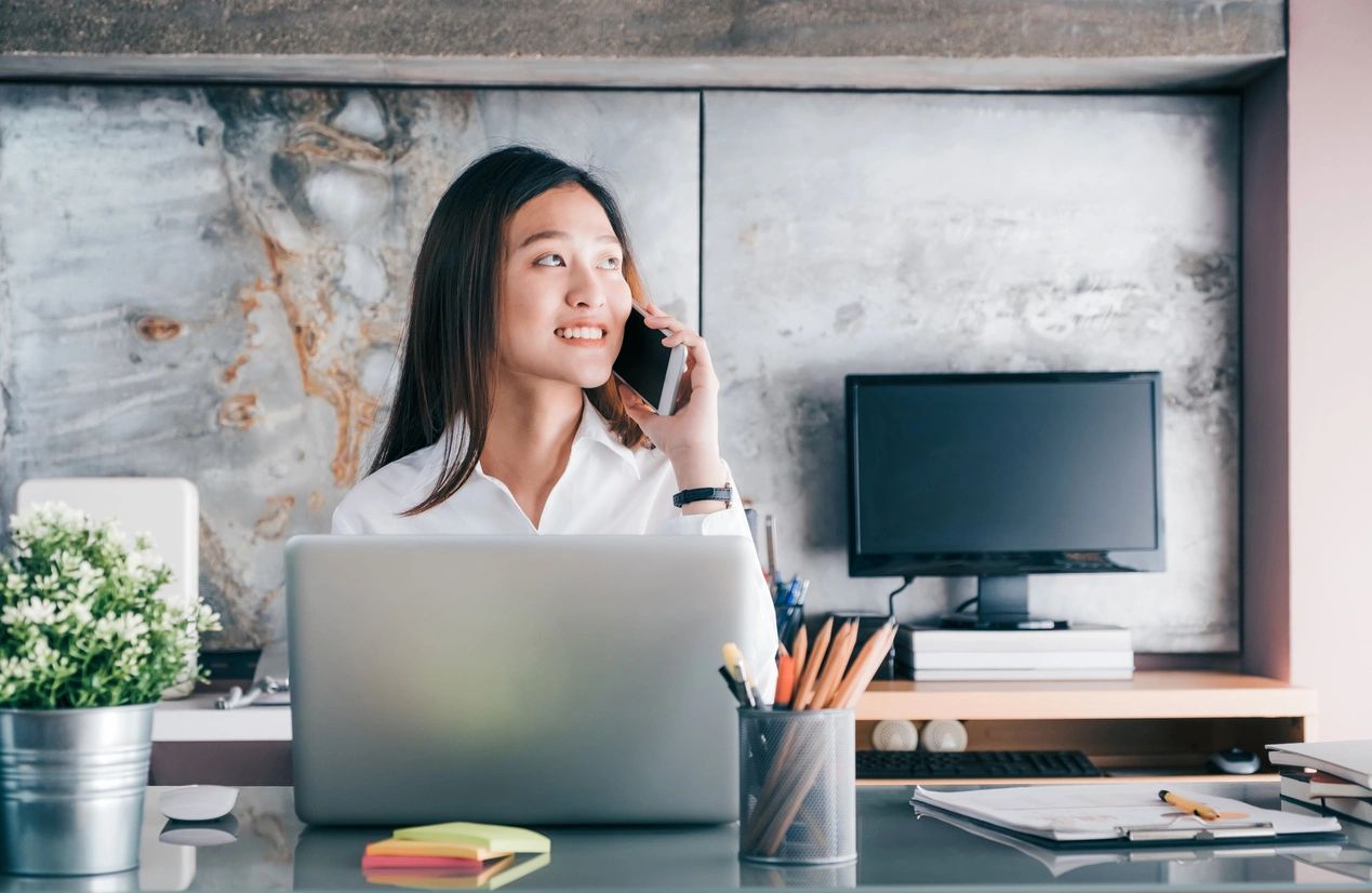 A woman talks on the phone while working on a laptop at her desk.